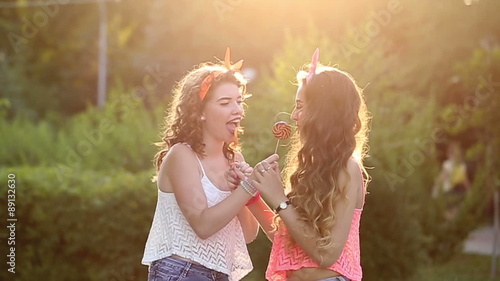 Cheerful friends walking in a summer park. Girl licking lollipop from each other. The concept of true friendship.