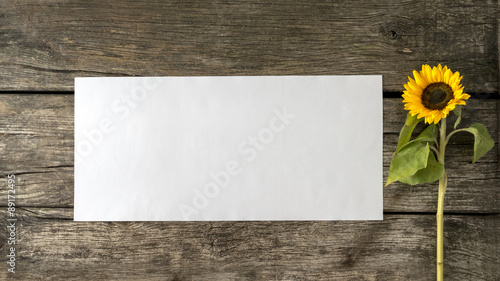 Fototapeta Naklejka Na Ścianę i Meble -  Blank white card or sheet of  paper with copyspace for your text on old textured rustic wooden planks and beautiful blooming sunflower on the right side.