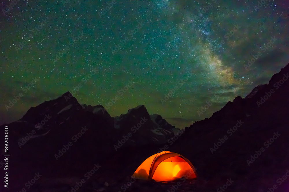 Night mountain landscape with illuminated tent.
Silhouettes of snowy mountain peaks and edges night sky with many stars and milky way on background illuminated orange tent on foreground