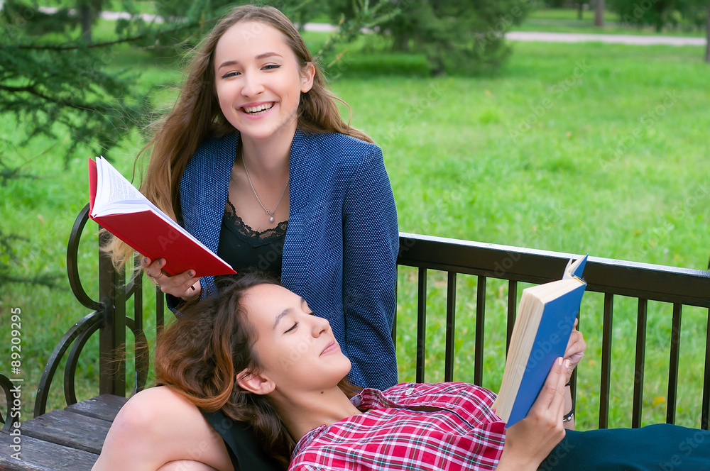 Fototapeta premium Two teenage girls reading books on bench in city park