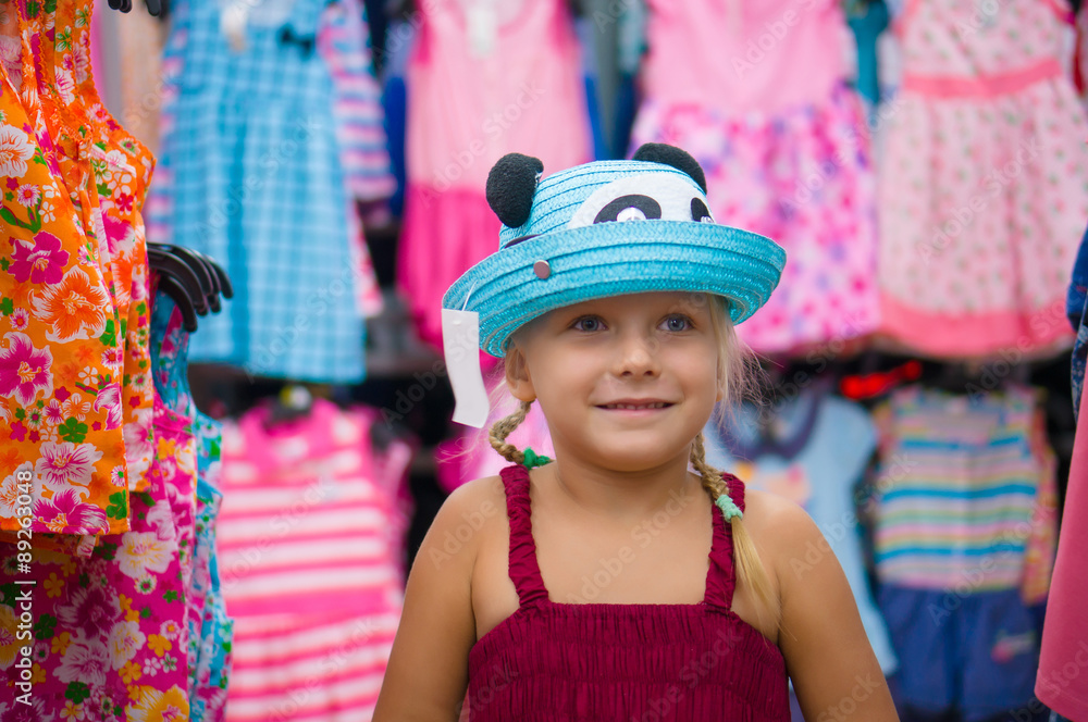 Adorable girl selects blue hat among beautiful baby girl dresses