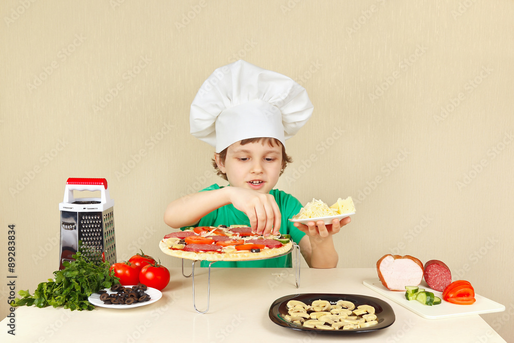 Young smiling boy in chefs hat puts a grated cheese on the pizza crust Young smiling boy in chefs hat puts a grated cheese on the pizza crust