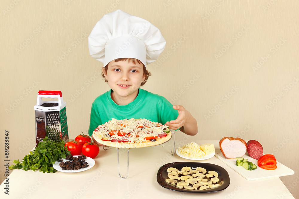 Young funny boy in chefs hat shows how to cook a pizza Young funny boy in chefs hat shows how to cook a pizza