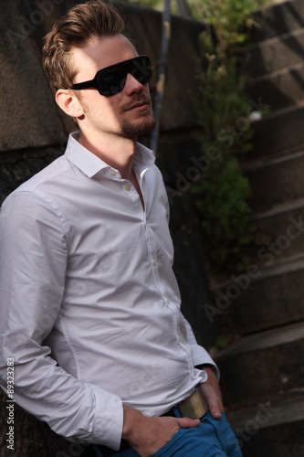 A young and attractive man in his 20s standing outside on a set of steps, wearing a white shirt, blue jeans and sunglasses, looking away from camera.