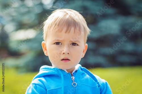 Closeup portrait of cute Caucasian boy with blond hair and dark brown eyes with funny face expression in blue hoodie outside in park on summer day, backlit with sun, rim light of his figure