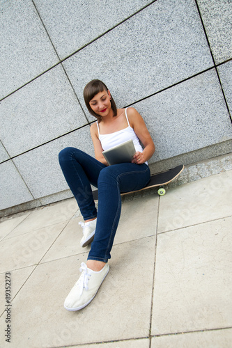 Young girl siting on skateboard in the cty with tablet by the wa © Wisiel