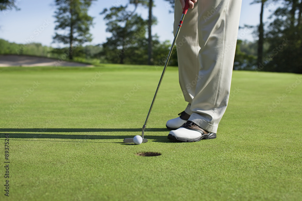 Obraz premium Golfer taps in with putter on green with trees near a lake