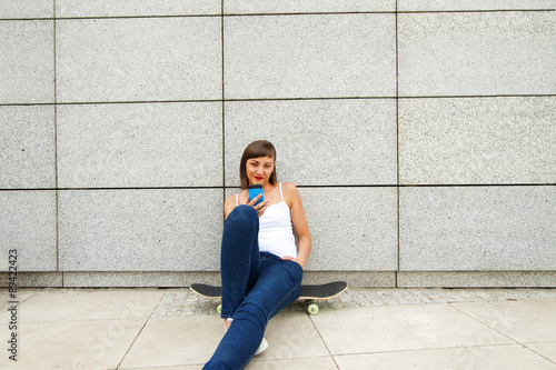Young girl siting on skateboard in the city with phone by the wa