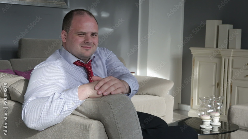 businessman sitting in living room on couch and smiles and nods at camera businessman sitting in living room on couch and smiles and nods at camera