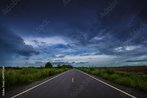 Asphalt road through the green field and clouds on dark sky in 