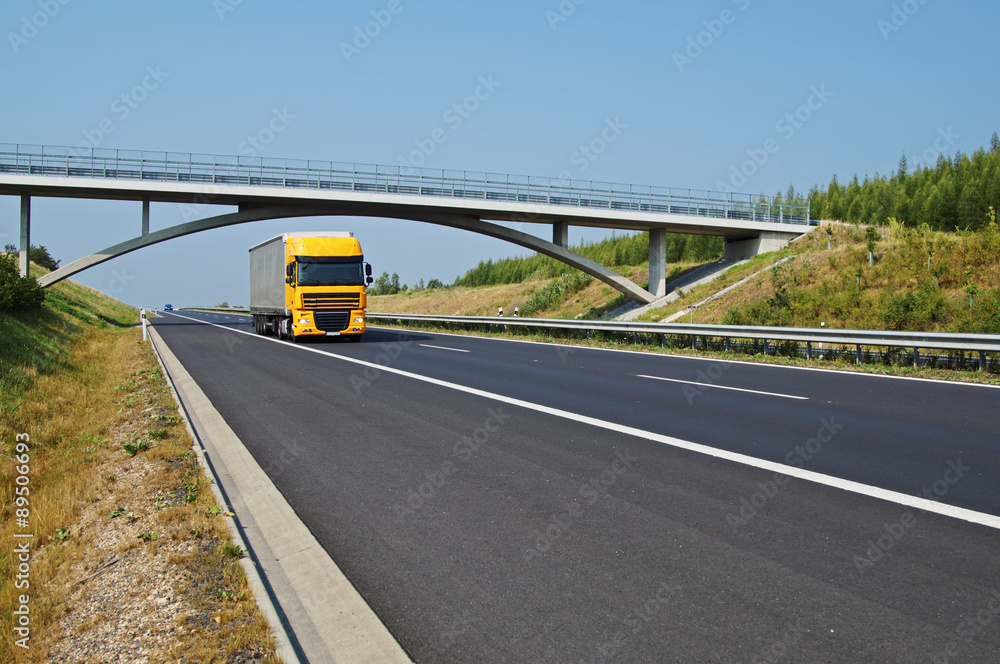 Yellow truck on highway goes under a concrete bridge in the countryside. Sunny day with blue sky.