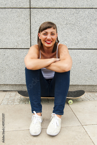 Young modern girl sit on skateboard by the wall in the city.
