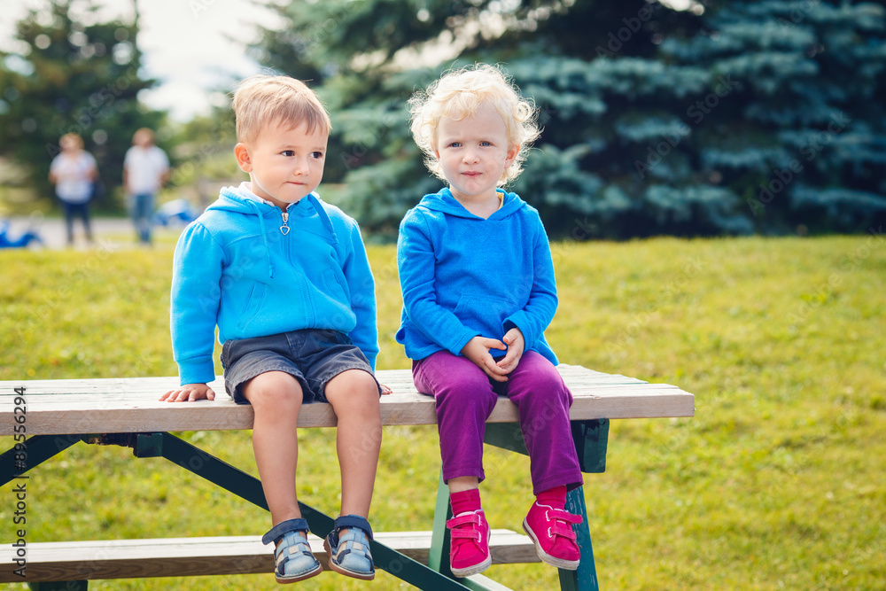Fototapeta premium Portraits of Caucasian cute boy and girl friends sitting together in park outside on summer day, backlit with sun from behind