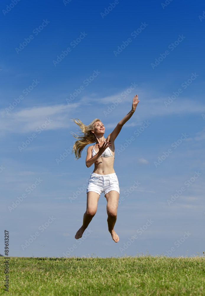 The happy woman in white bikini and shorts jumps in a summer green field against the blue sky..