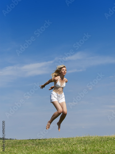 The happy woman in white bikini and shorts jumps in a summer green field against the blue sky.. © Konstantin Kulikov