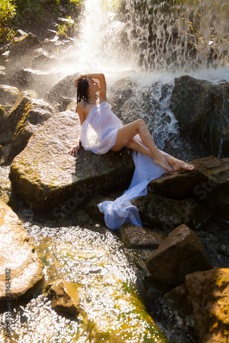 Naked girl sitting on a rock by a waterfall.