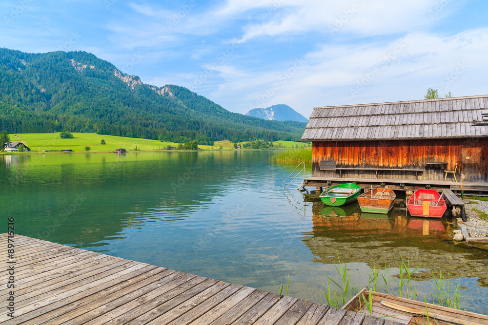 Wooden jetty and fishing boats with wooden houses on shore of Weissensee lake in summer landscape of Carinthia land, Austria