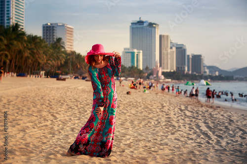 slim girl in long and big hat on beach against city sea