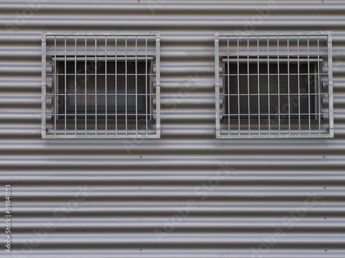 Two barred windows on a grey facade made of corrugated iron.