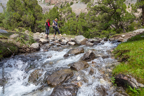 Two hikers crossing fast flowing river People going across mountain creek with fast streaming water jumping on the rocks green meadow and forest along river