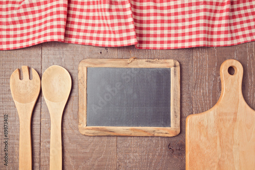Abstract wooden background with red checked tablecloth, chalkboard and kitchen utencils. View from above