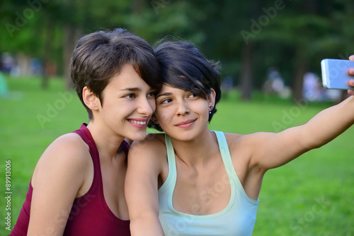 Two young short hair women taking photos with phone in a park.