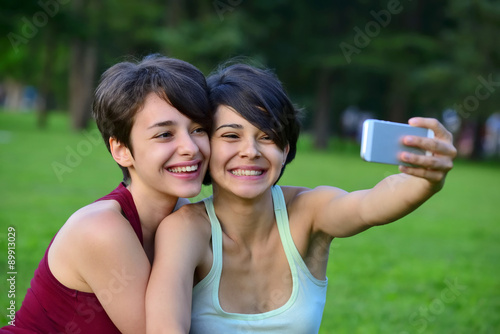 Two young short hair women taking photos with phone in a park.