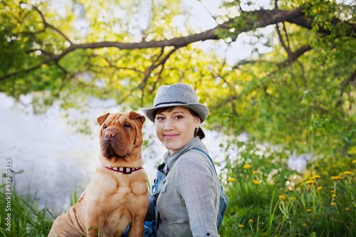 Obraz na plátně happy young woman in a hat with dog Shar Pei sitting in the field in sunset ligh