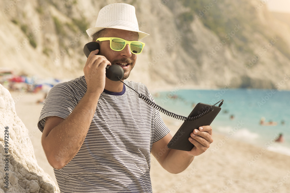 Young guy making a call from the beach by using digital tablet device and retro telephone handset 
