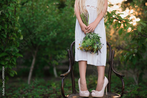 The little girl in high-heeled shoes large size in a white dress standing on a chair with a vintage bouquet in the hands of fashion, lifestyle, wedding