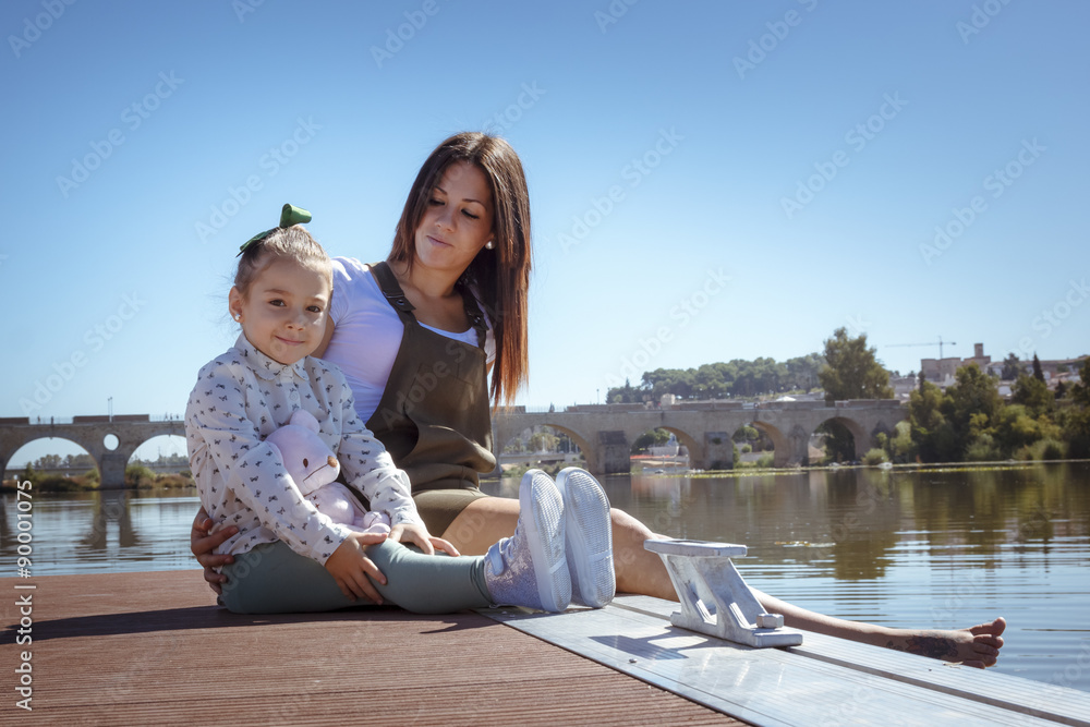 Madre e hija sentadas en el embarcadero. Una niña y su mama disfrutando del verano junto al rio. Madre joven soltera.