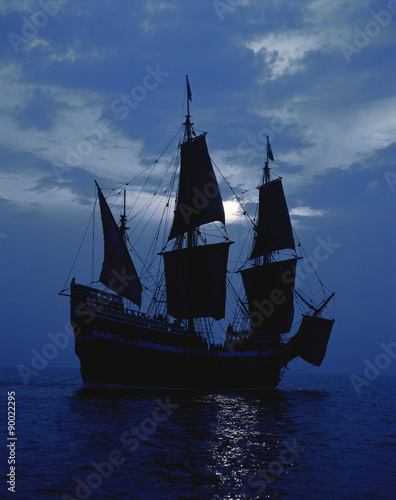 This is a replica of the ship Mayflower II. It demonstrates the first sailing in 1620 when the Pilgrims sailed to the New World. The ship shows full sails on a shimmering moonlit ocean against a dark blue night sky.