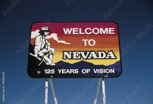 This is a road sign that says, Welcome to Nevada, 125 years of Vision. It is located on Interstate 15. It shows a miner with a pick in his hand, squatting next to the mountains against a red, orange and blue background. There is also a cloud in the sky.
