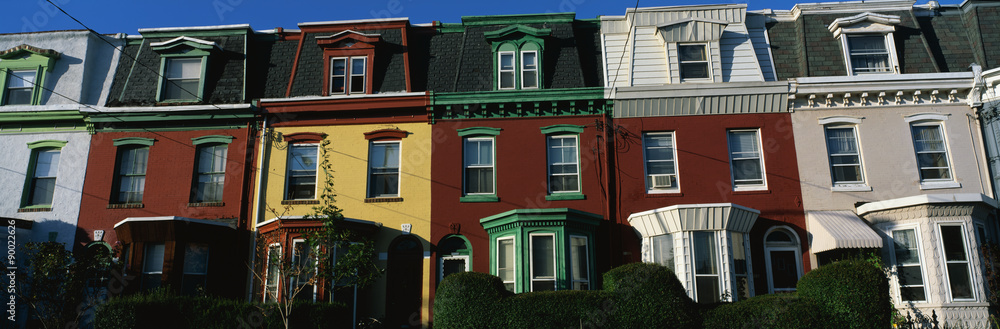 These are typical urban style row houses. They are all lined up next to each other with neatly trimmed bushes in front of them. They are colorfully painted in red, white or yellow paint. They all have a single window on the top floor with two windows on the second floor. They have bay windows on the first floor.