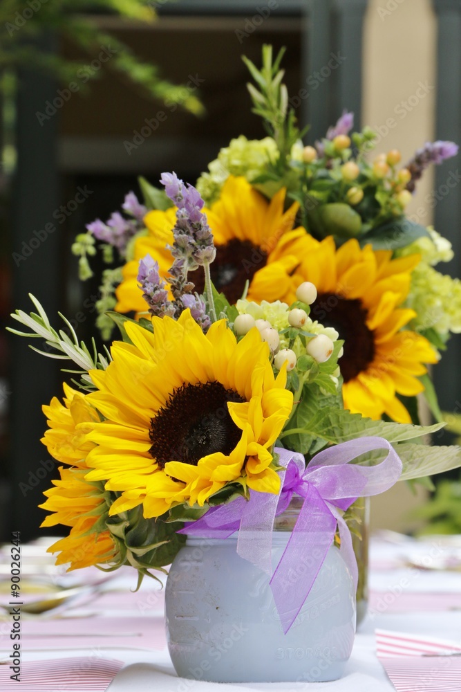 Fototapeta premium Objects: Sunflowers in a flower arrangement with a bow on a table with napkins, silverware and balloons in the background.