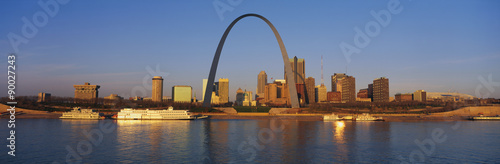 This is the skyline at sunrise. It is situated along the Mississippi River. There are riverboats on the water with the St. Louis Arch in clear view.