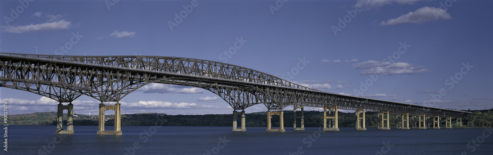 This is the Beacon Bridge which is over the Hudson River. It is a large steel bridge. The water and sky are blue.