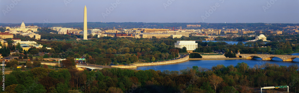 This is an aerial view of Washington, DC with the Jefferson Memorial, U.S. Capitol, Washington Monument, and Lincoln Memorial. The Potomac River runs through the center with the Key Bridge at right at sunset. There is green foliage of the spring as well.