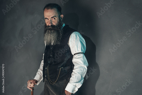 Vintage beard man in 1900 style fashion with cane against grey w