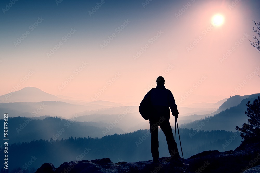 Silhouette of tourist with poles in hand. Hiker with sporty backpack stand on rocky view point above misty valley. Sunny morning in rocky mountains.