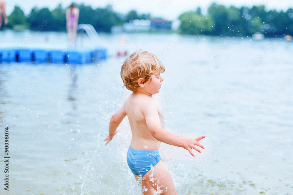 Little blond kid boy having fun with splashing in a lake, outdoo