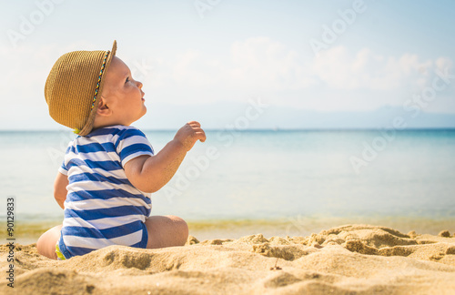 Baby boy with hat sitting on the beach.