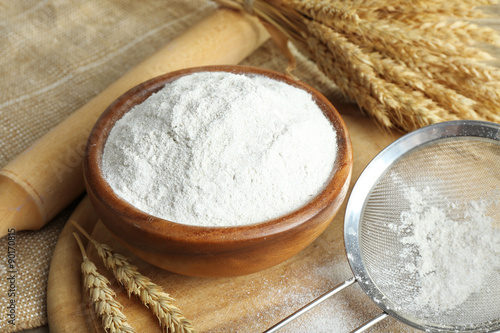 Whole flour in bowl with wheat ears on wooden table, closeup