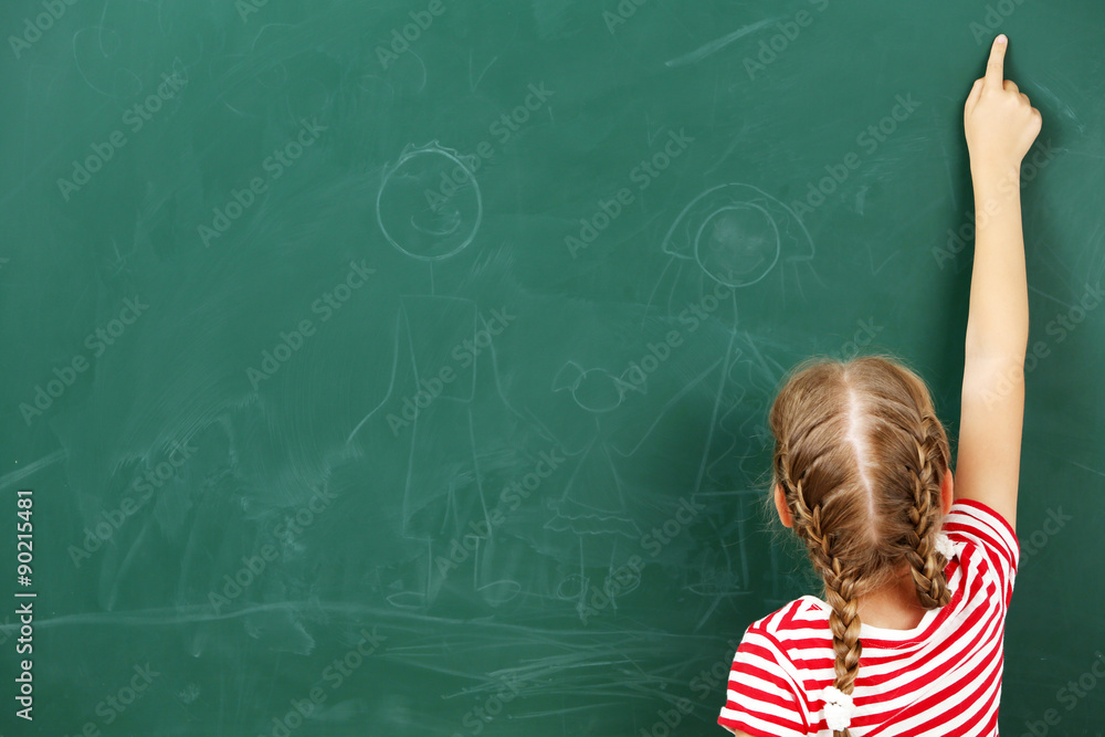 Little girl pointing at something at black chalkboard in classroom