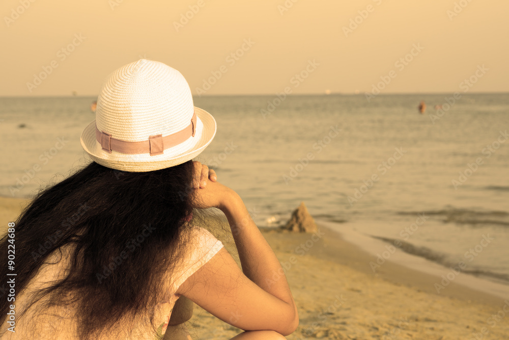 Girl brunette in white hat looking at sea while sitting on beach warm filtered