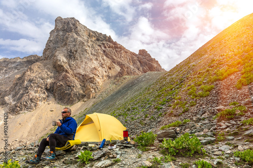 Lunch of alpine climber Hiker sitting aside yellow camping tent and having lunch stove and cooking gear mountain landscape shining sun sunbeams on background