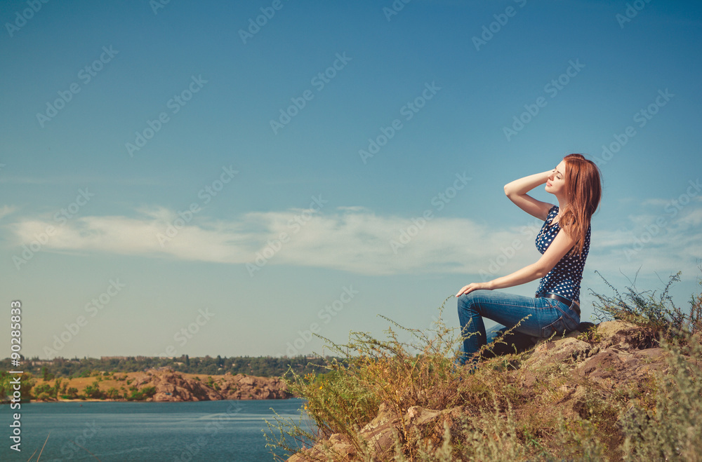 Beautiful young woman sitting on the cliff above the river, meditating with her eyes closed
