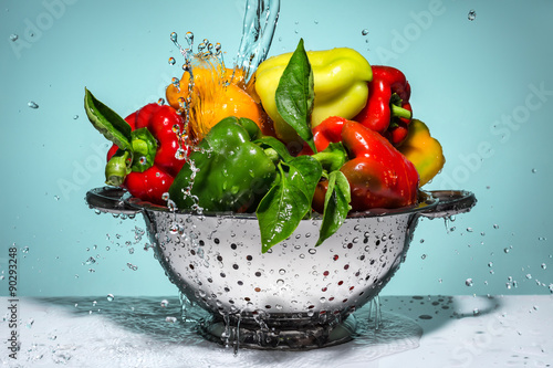 Peppers of different colors in a colander under running water.