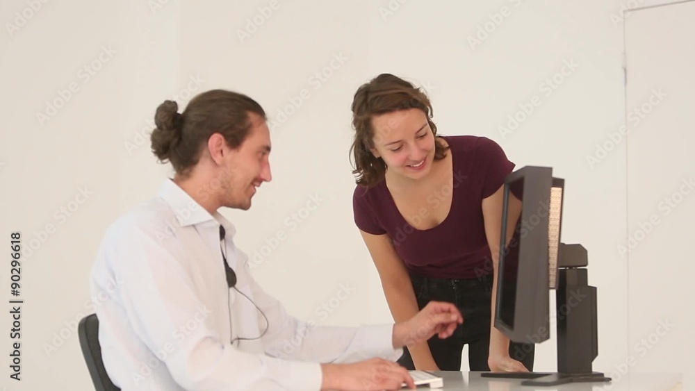 Two young colleagues, a boy sitting and a girl bowing next to his desk, talking and pointing at a computer monitor Two young colleagues, a boy sitting and a girl bowing next to his desk, talking and pointing at a computer monitor