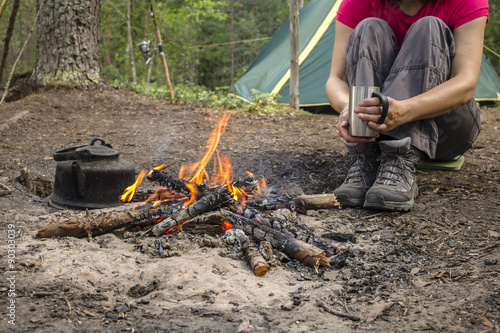 Girl in a red T-shirt sitting with cup while camping near the bonfire close to tent is heated and drink hot tea